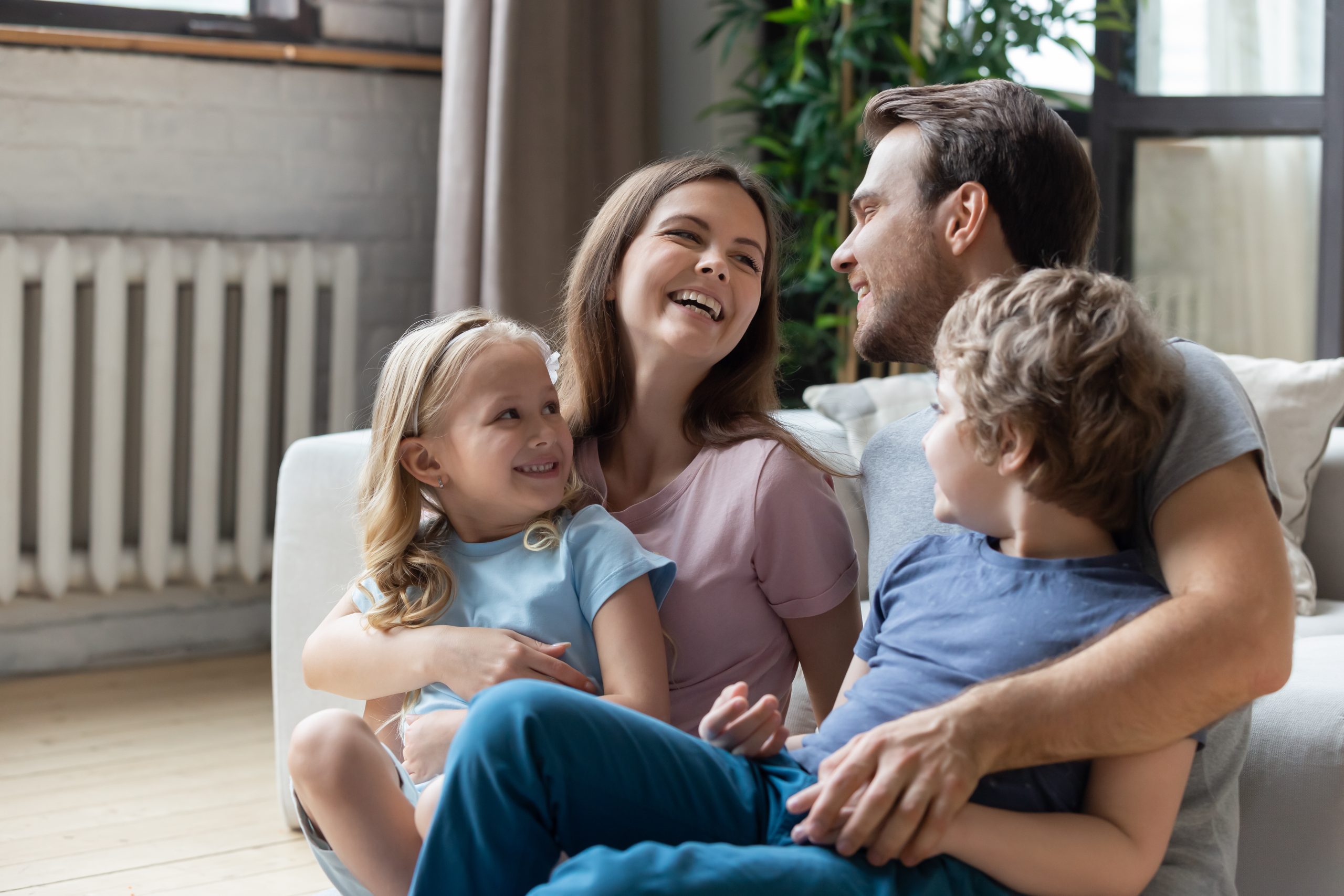 Happy parents with little son and daughter sitting on warm floor in living room, laughing mother and father looking at each other, hugging kids, having fun at home, family enjoying tender moment